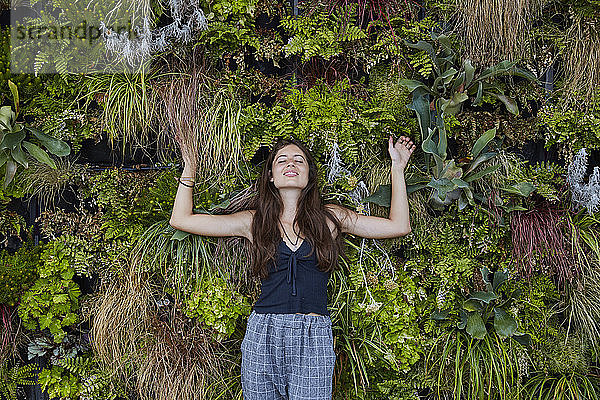 Porträt einer lächelnden jungen Frau mit geschlossenen Augen vor einer Pflanzenwand
