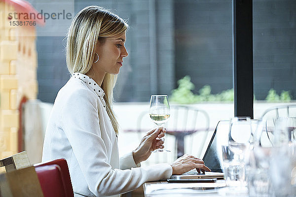 Geschäftsfrau benutzt Laptop in einem Restaurant  während sie Weißwein trinkt