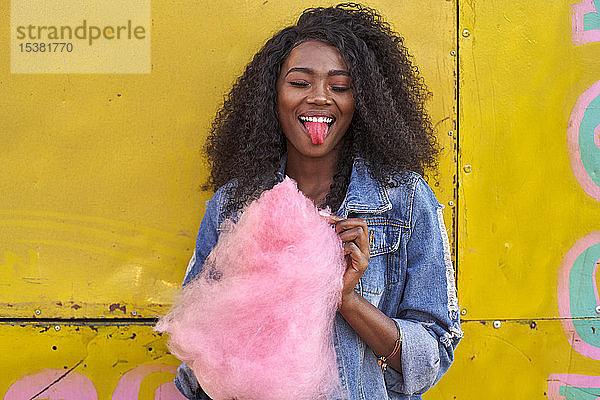Porträt einer lächelnden jungen Frau mit herausgestreckter Zunge aus rosa Zuckerwatte