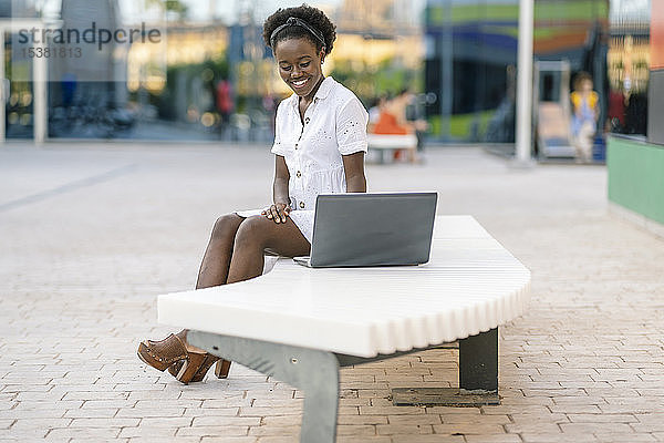 Porträt einer lächelnden jungen Frau  die auf einer Bank sitzt und auf einen Laptop schaut
