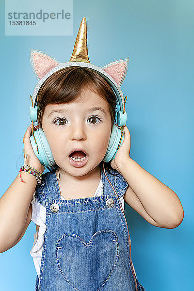 Porträt eines süßen kleinen Mädchens  das mit einhornförmigen Kopfhörern auf blauem Hintergrund Musik hört und singt