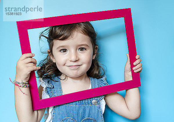 Porträt eines niedlichen kleinen Mädchens  das einen Bilderrahmen auf blauem Hintergrund hält