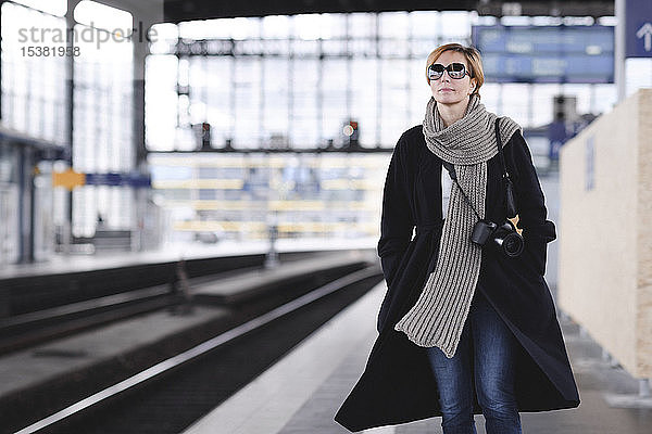 Reife Frau mit Kamera in schwarzem Mantel und großem Wollschal wartet am Bahnsteig