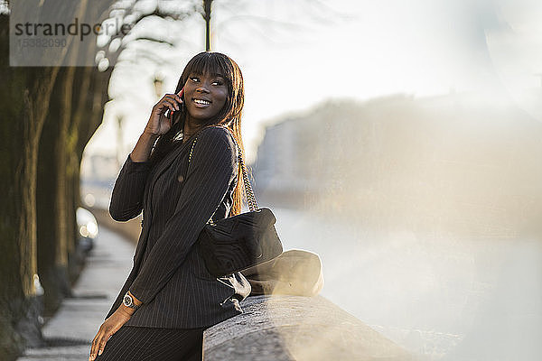 Lächelnde Geschäftsfrau steht am Flussufer und benutzt ihr Smartphone
