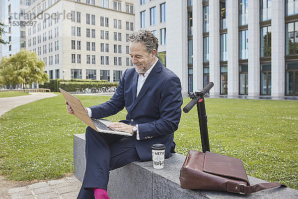 Glücklicher reifer Geschäftsmann  der in der Stadt mit einem Laptop an einer Wand sitzt