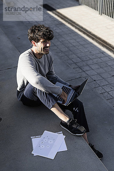 Lächelnder Mann sitzt auf einer Außentreppe mit Papieren und Laptop