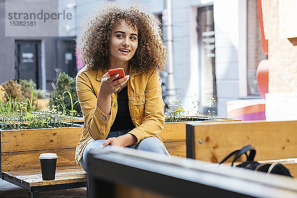 Porträt einer Teenagerin am Telefon  die auf einer Bank sitzt und Kaffee zum Mitnehmen