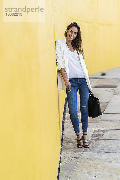 Porträt einer lächelnden Frau  die an einer gelben Wand steht und eine Handtasche hält