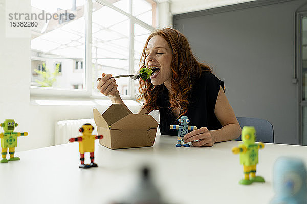 Porträt einer rothaarigen Geschäftsfrau in einem Loft mit Miniaturrobotern auf dem Schreibtisch  die gesundes Essen zum Mitnehmen essen
