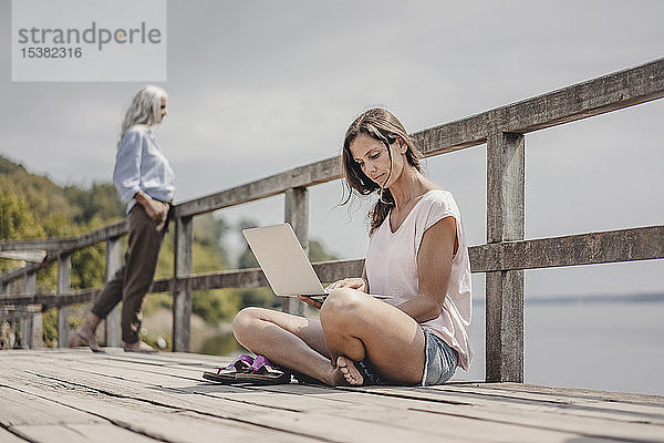 Frau auf Holzbrücke sitzend  mit Laptop  Mutter im Hintergrund stehend