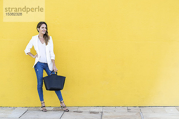 Lächelnde Frau steht an einer gelben Wand und hält eine Handtasche
