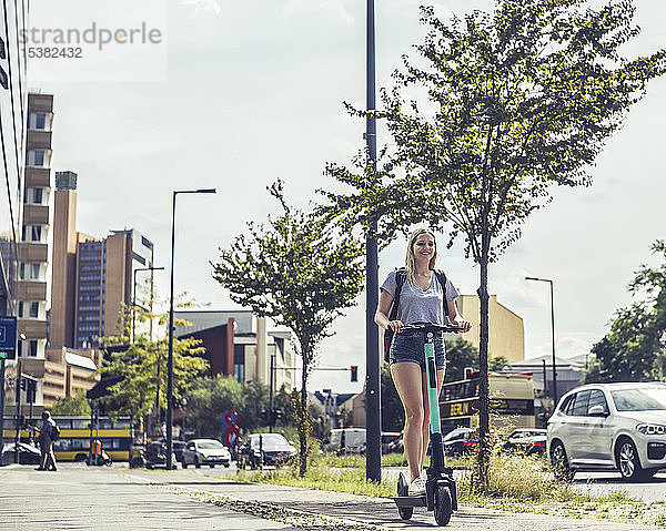 Lächelnde junge Frau fährt E-Scooter auf dem Fahrradweg  Berlin  Deutschland