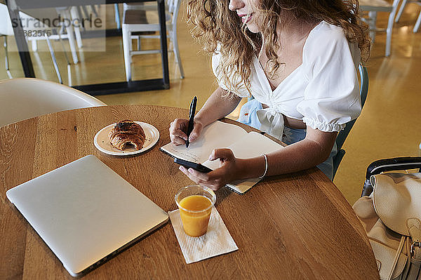 Junge Frau in einem Café  die ein Smartphone benutzt und sich während des Frühstücks Notizen macht