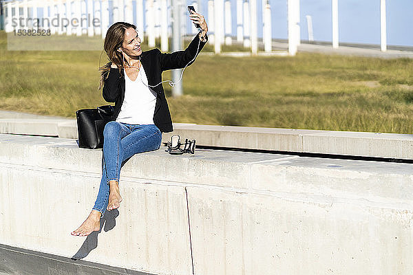 Lächelnde Geschäftsfrau sitzt mit Smartphone und Kopfhörern an der Wand