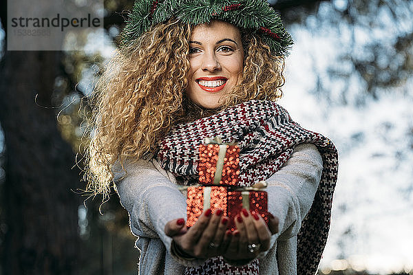 Porträt einer lächelnden jungen Frau mit Weihnachtskranz auf dem Kopf  die ein Weihnachtsgeschenk überreicht