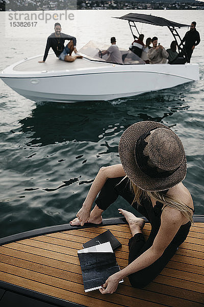 Frau mit einem Buch auf einem Boot  die ein Boot mit jungen Männern betrachtet