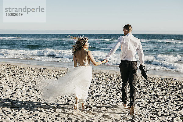 Rückansicht von Braut und Bräutigam beim Laufen am Strand