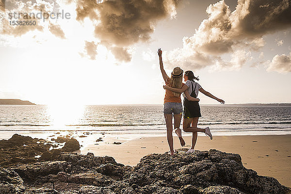 Zwei Freundinnen stehen am felsigen Strand  winken bei Sonnenuntergang  Rückansicht