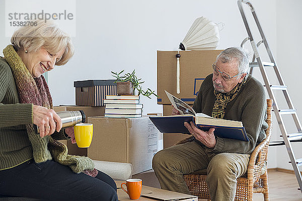 Älteres Ehepaar mit Fotoalbum und Heißgetränk umgeben von Pappkartons in einem leeren Raum