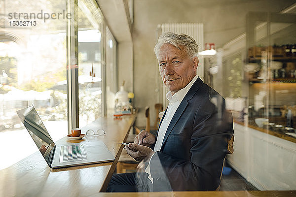 Porträt eines älteren Geschäftsmannes mit Handy und Laptop in einem Café