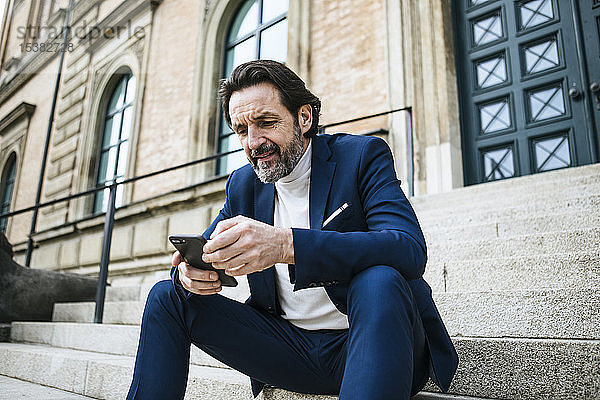 Porträt eines bärtigen  reifen Geschäftsmannes  der auf einer Treppe sitzt und auf sein Handy schaut