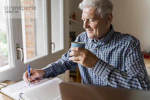 Älterer Mann sitzt zu Hause vor dem Laptop und macht sich Notizen