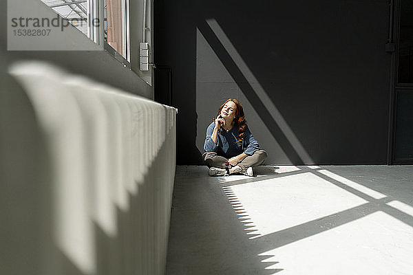 Rothaarige Frau sitzt auf dem Boden eines Dachbodens und genießt das Sonnenlicht