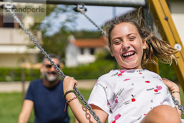 Porträt eines glücklichen Mädchens auf einer Schaukel mit dem Vater im Hintergrund