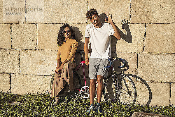 Junges Paar mit Fahrrad  an Steinmauer gelehnt  Handzeichen machen