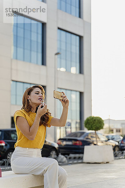 Geschäftsfrau sitzt im Stadtzentrum und benutzt ein Smartphone  um Lippenstift aufzutragen