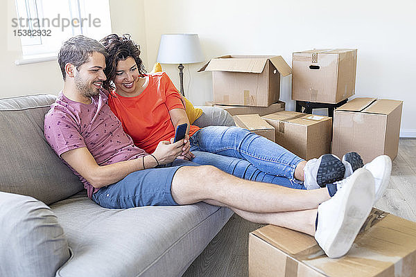 Glückliches Paar benutzt Handy auf der Couch im neuen Zuhause