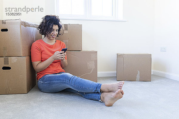 Frau sitzt in neuer Wohnung auf dem Boden und benutzt Mobiltelefon