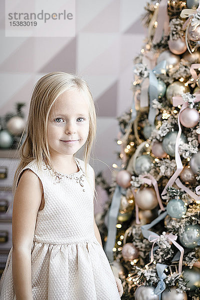 Porträt eines lächelnden kleinen Mädchens  das vor einem beleuchteten Weihnachtsbaum steht