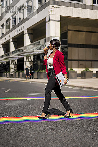 Geschäftsfrau  die Kaffee zum Mitnehmen trinkt  während sie auf LGBT-Streifen über die Straße geht  London  UK