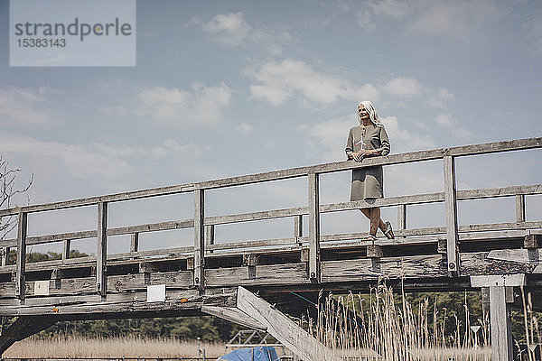 Ältere Frau steht auf Holzbrücke und hält Modell einer Windkraftanlage