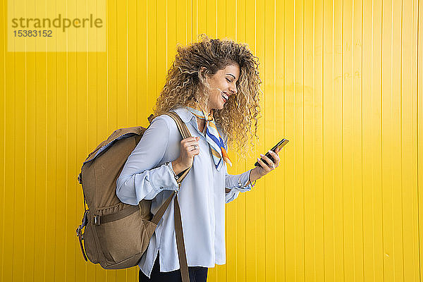 Lächelnde junge Frau mit Rucksack vor gelbem Hintergrund schaut auf Handy