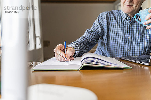 Älterer Mann sitzt zu Hause vor dem Laptop und macht sich Notizen