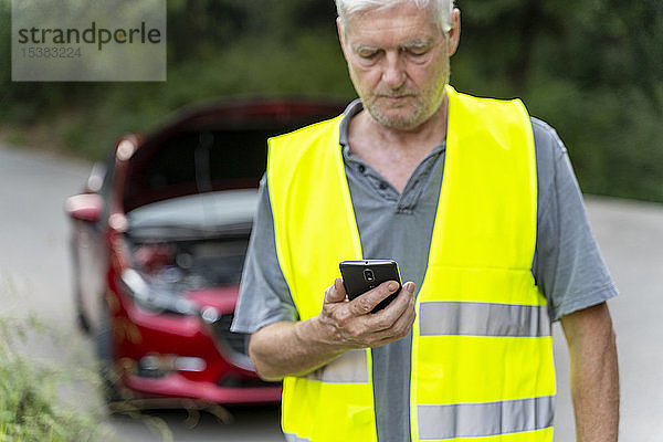 Älterer Mann steht an seinem kaputten Auto  trägt eine Sicherheitsweste und benutzt sein Smartphone