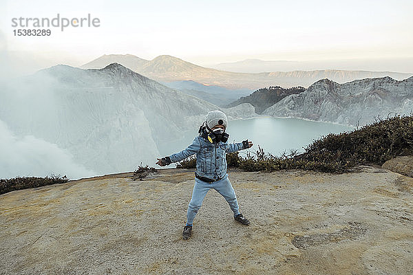 Junge mit Atemschutzmasken steht mit ausgestreckten Armen am Rande des Vulkans Ijen  Java  Indonesien