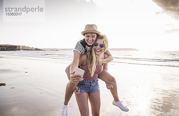 Zwei Freundinnen  die sich am Strand vergnügen  sich gegenseitig huckepack tragen  sich selbst ein Smartphone nehmen