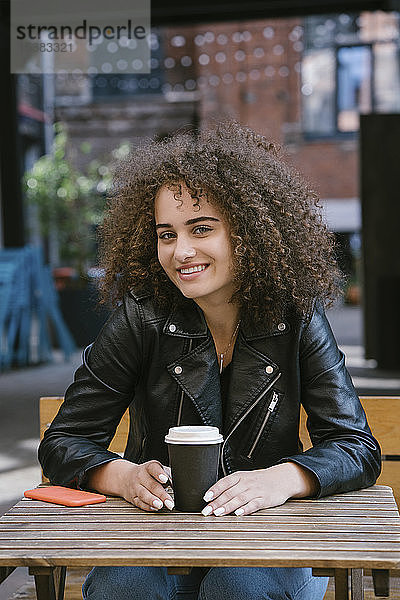 Porträt einer lächelnden Teenagerin  die auf einer Bank sitzt  mit Kaffee zum Mitnehmen und Mobiltelefon