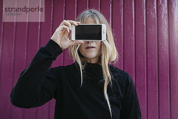 Porträt einer blonden jungen Frau  die mit einem Smartphone die Augen verdeckt