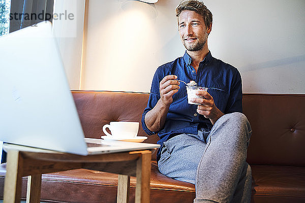 Junger Geschäftsmann isst und benutzt Laptop in einem Café