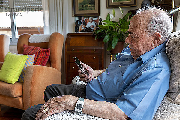 Älterer Mann  der zu Hause ein Mobiltelefon benutzt und ein intelligentes Notfall-Alarmarmband um das Handgelenk trägt