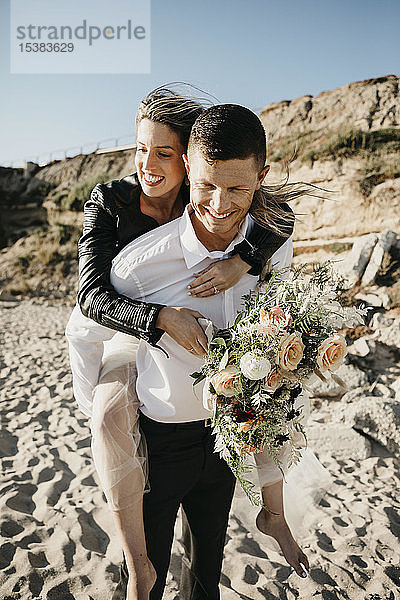 Glücklicher Bräutigam trägt Braut huckepack am Strand