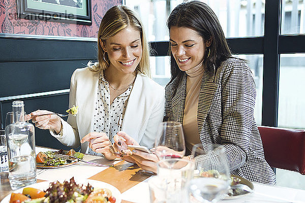 Zwei glückliche Geschäftsfrauen in einem Restaurant  die sich gemeinsam ein Smartphone anschauen