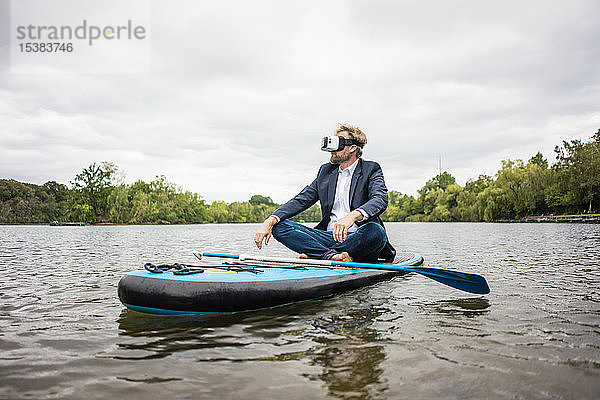 Geschäftsmann mit VR-Brille sitzt auf einem SUP-Brett auf einem See