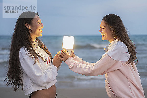 Zwei glückliche Freundinnen  die in der Abenddämmerung am Strand ein Führungslicht halten