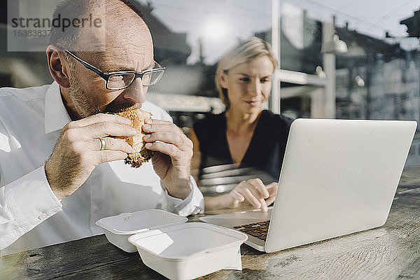 Geschäftsmann isst Hamburger im Café  während ein Kollege seinen Laptop benutzt