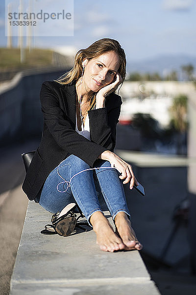 Porträt einer selbstbewussten Geschäftsfrau  die mit Smartphone und Kopfhörern an einer Wand sitzt